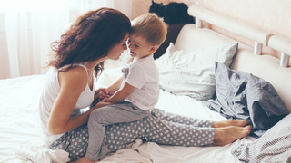 A woman and a young child sitting together on a bed in a bedroom setting. The woman has dark hair and is wearing a white tank top and gray polka-dot pajama pants, while the child wears a white t-shirt and gray pants.
