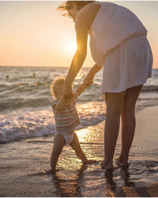 A woman in a white sleeveless dress and a toddler in a striped shirt standing in shallow ocean water at sunset, holding hands on a beach.